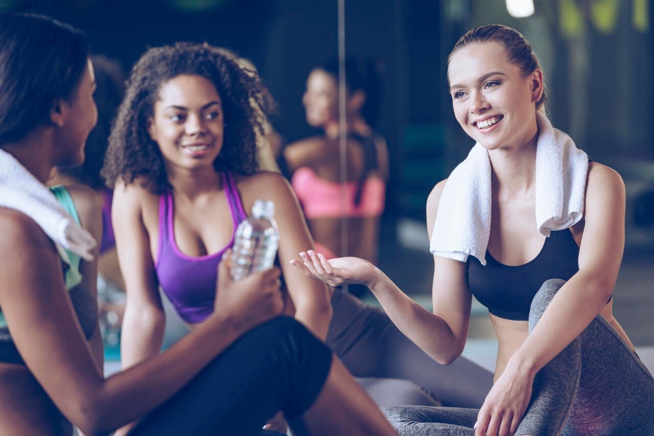 Beautiful young women in sportswear discussing something with smile while sitting on exercise mat at gym