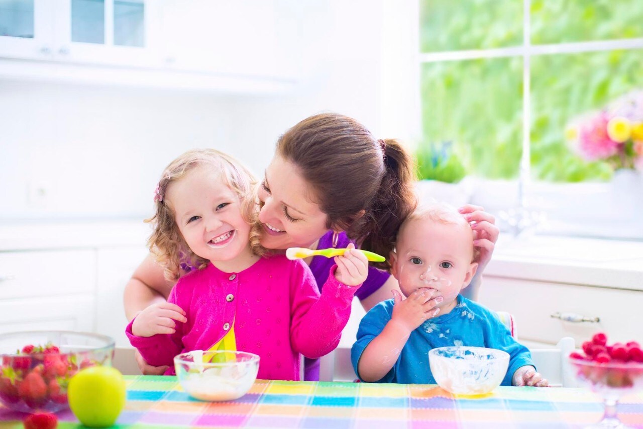Happy young family, mother with two children, adorable toddler girl and funny messy baby boy having healthy breakfast eating fruit and dairy, sitting in a white sunny kitchen with window