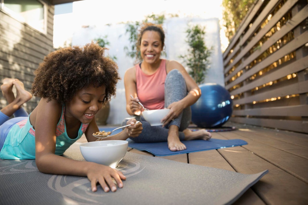 African American mother and daughter eating cereal on yoga mats on deck